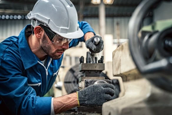A male worker in a blue jumpsuit and white hardhat operates a lathe.